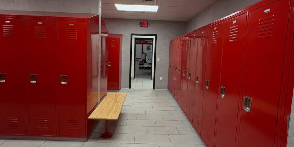 Row of red metal lockers in a grey locker room with a wooden bench.