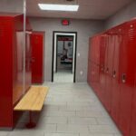 Row of red metal lockers in a grey locker room with a wooden bench.