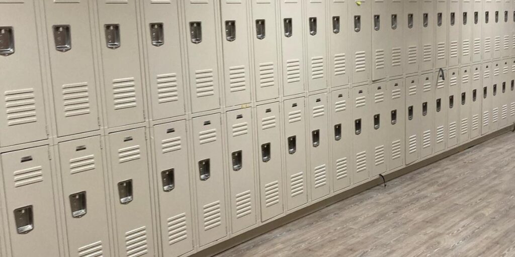 A long row of tan metal lockers in two tiers along a wall with wood-look flooring.