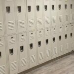 A long row of tan metal lockers in two tiers along a wall with wood-look flooring.