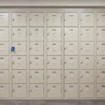 High-resolution row of cream-colored metal lockers in a clean, modern indoor hallway with blue locks, tiled flooring, and a countertop cabinet.